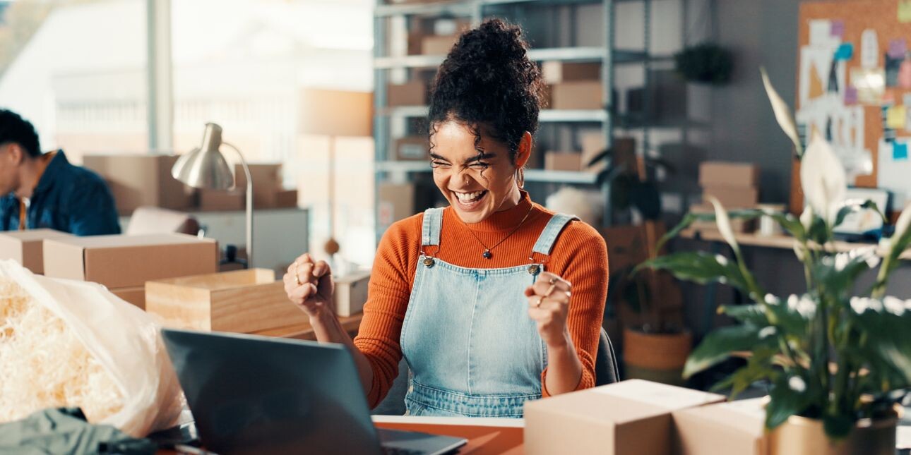 A happy woman raising her arms in delight while sat at her laptop.